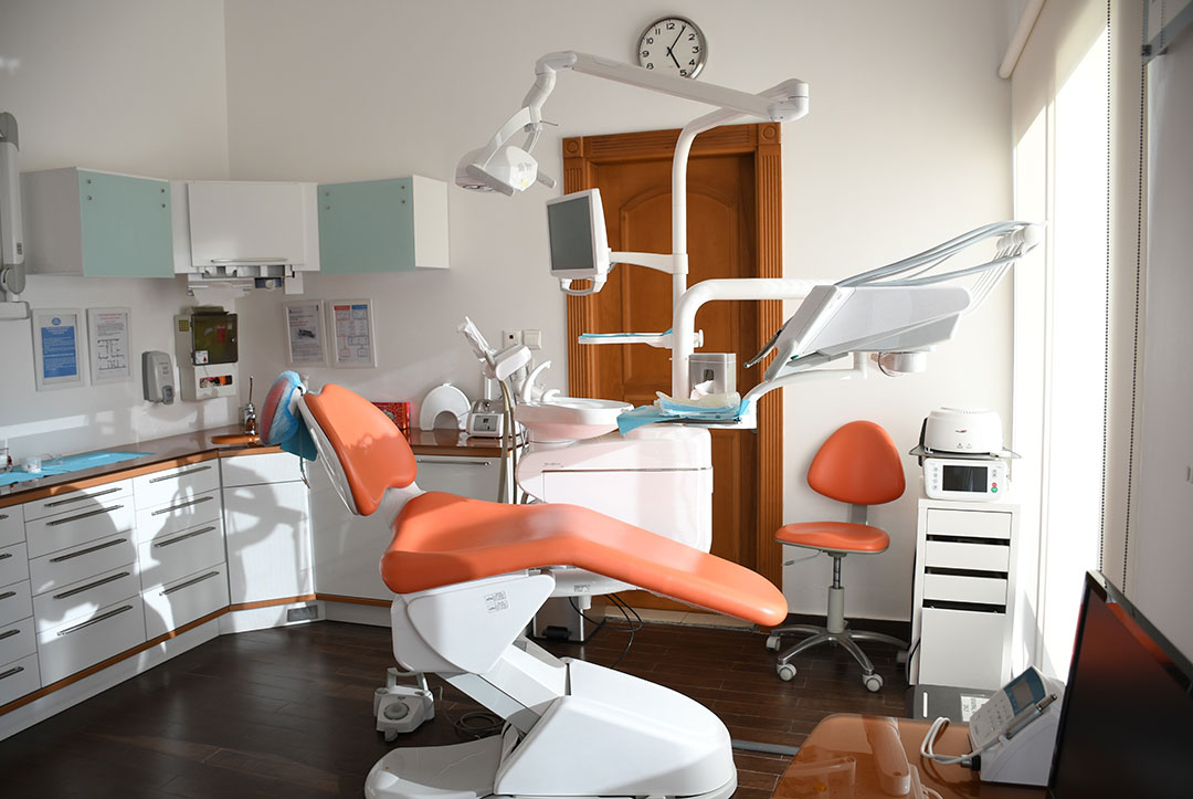 Sauberes Behandlungszimmer eines Zahnarztes mir weißem Inventar und orangfarbenen Behandlungsstuhl.